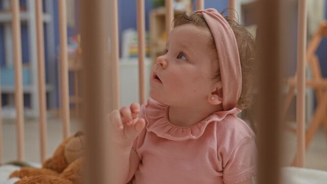 Girl in a pink headband reaches arm for a teddy bear through crib slats inside a nursery building; curiosity comfort.