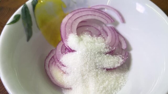 A spoonful of granulated sugar above sliced red onion in a ceramic bowl on a wooden cutting board during food preparation.