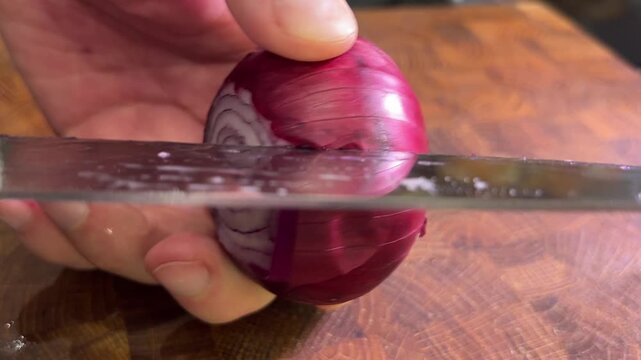 Closeup of hands holding and cutting a red onion with a kitchen knife on a textured wooden cutting board.