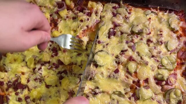 A knife and fork cut into homemade pizza covered with melted cheese, sausage, chicken, pineapple, and pickle slices.