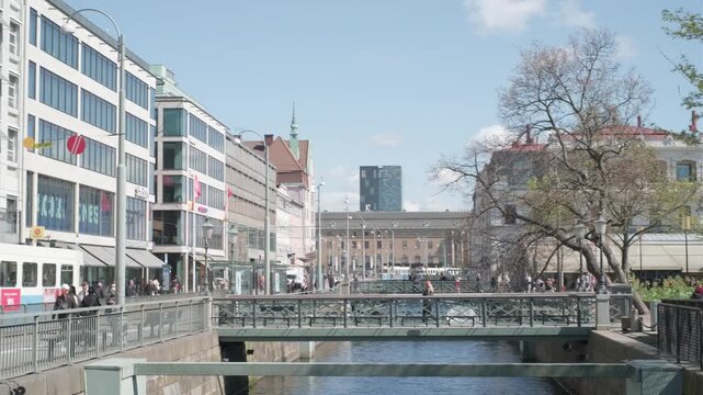 Gothenburg, Sweden - May 2 2023: Pedestrians cross a bridge over a canal in central Gothenburg, with cafes, shops, a tree-lined promenade and Gustaf Adolfs Torg visible in the background under a partl