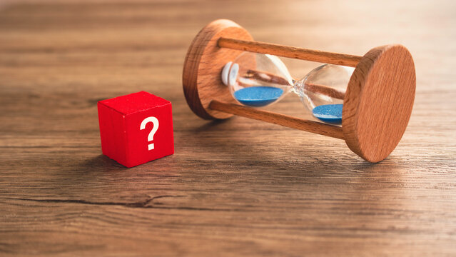 Red question mark cube beside lying hourglass on wooden table financial or career time uncertainty concept