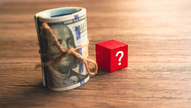 Vertical roll of hundred dollar bills tied with rope beside red question mark cube financial planning decision concept