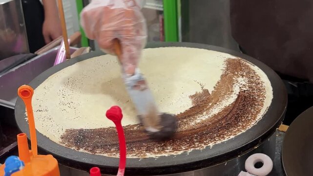 Close up 4K footage of a gloved vendor spreading rich chocolate filling across a thin crepe cooking on a large circular griddle at a food stall.