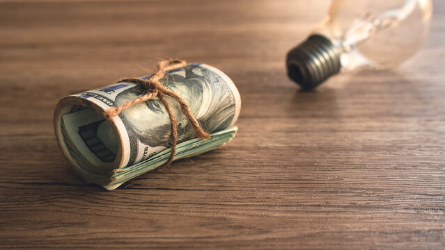 Rolled US Dollar Banknotes Tied with String Beside Incandescent Light Bulb on Wooden Desk Concept for Investment Ideas, Utility Bills, and Business Innovation