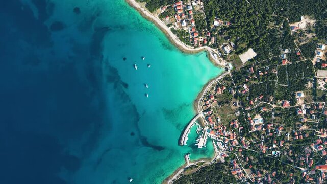 A high-angle top-down aerial view of a picturesque turquoise bay and harbor on Silba Island, Croatia, showing moored boats, lush green vegetation, and vibrant red-roofed houses along the crystal-clear