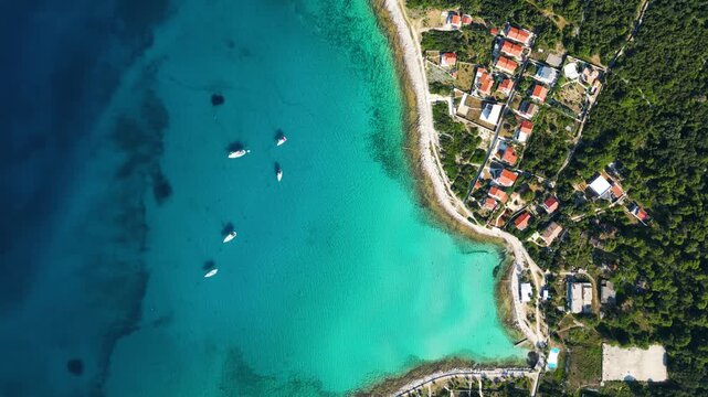 A high-angle top-down aerial view of a picturesque turquoise bay and harbor on Silba Island, Croatia, showing moored boats, lush green vegetation, and vibrant red-roofed houses along the crystal-clear