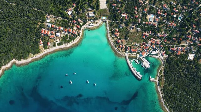 A high-angle top-down aerial view of a picturesque turquoise bay and harbor on Silba Island, Croatia, showing moored boats, lush green vegetation, and vibrant red-roofed houses along the crystal-clear