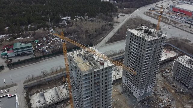 Aerial view of three high-rise buildings under construction. Yellow cranes loom above unfinished concrete structures. Workers move between floors, busy with building tasks