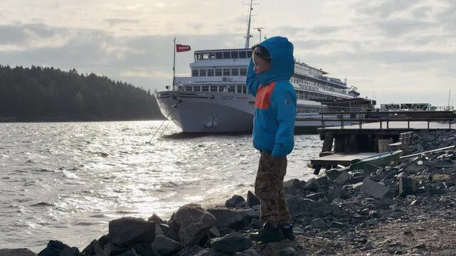 A curious boy plays with rocks on a stony shore. A cruise ship rests peacefully near the dock on Lake Ladoga, Karelia. He feels excitement and wonder during this peaceful adventure.