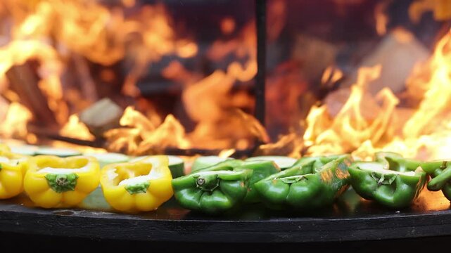 Colorful bell peppers and zucchini grilling on round barbecue grill with smoke. Fresh vegetables cooking over charcoal fire. Street food and outdoor barbecue concept.