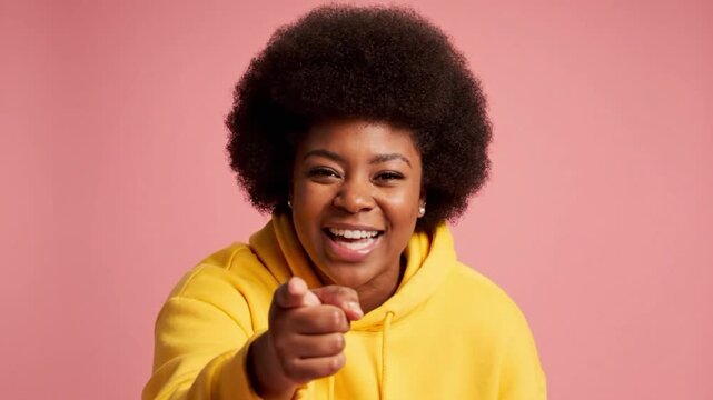 Girl with an afro hairstyle in a yellow hoodie laughs and interacts with the camera in a pink background setting