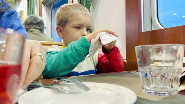 A young boy carefully wipes his hands with a napkin at a dining table. He feels calm and content inside a cozy cruise ship on Lake Ladoga. Peaceful feelings of care fill the room.