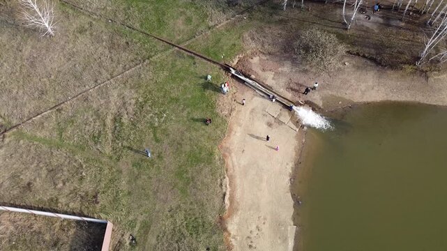 Aerial view shows a large pipe discharging water into a lake. People stand near the pipe, observing the flow. The scene captures an industrial or environmental process. It might represent wastewater