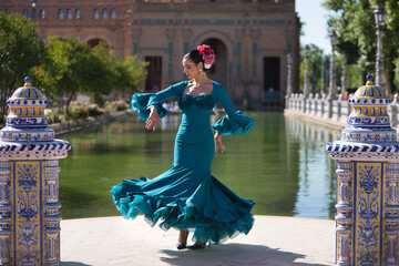 Young, brunette, Andalusian woman, beautiful, wearing a green flamenco dress and flowers in her hair, dancing flamenco, with the canal in the background, in Spain Square in Seville. © Manuel