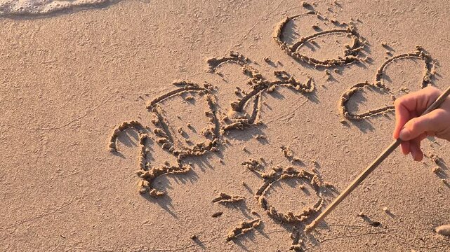Beach vacation drawing, a hand uses a stick to etch a sun into the sand near the ocean. High-angle static shot.