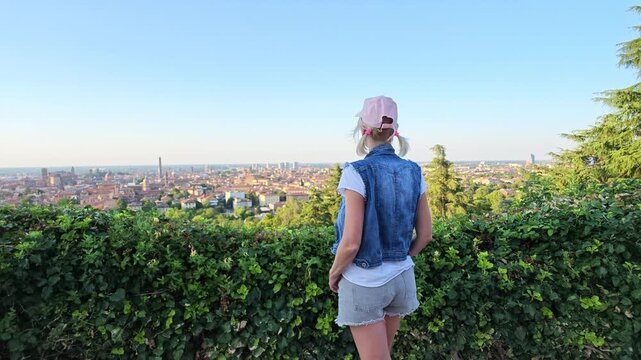 Woman standing on a hill viewing panoramic cityscape of Bologna, Italy, with historic architecture, including famous Two Towers, and Basilica Saint Petronius, dedicated to patron saint of Bologna.