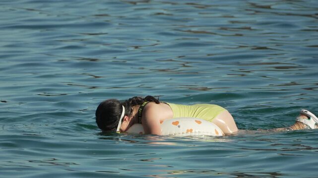 Floating woman snorkeling with yellow goggles on heart-patterned inflatable ring in sea. Static medium shot.