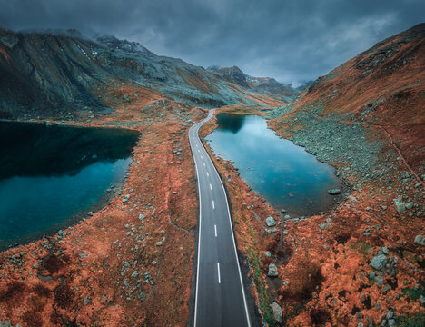 Aerial view of mountain road between two lakes in alpine valley in cloudy autumn day in Switzerland. Top drone view of empty road in swiss mountain pass, orange grass, ponds, overcast sky in fall
