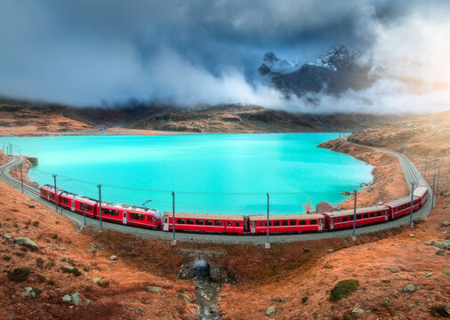 Aerial view of red Bernina Express train curving along the turquoise lake Bianco in the Swiss Alps, rocky hills and dramatic cloudy sky in autumn alpine mountain landscape. Top view of train in fall