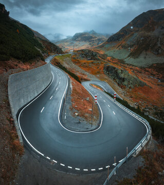 Aerial view of winding wet mountain road in alpine valley in rainy autumn day in Switzerland. Top drone view of empty road in swiss mountain pass, orange grass, stones, overcast sky in fall. Landscape