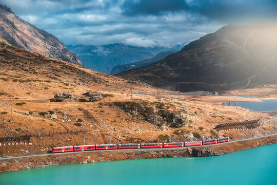 Aerial view of modern red train near azure mountain lake and dramatic cloudy sky at sunset in autumn. Bernina Express, Switzerland. Top view of train in Alps, winding railroad, orange hills in fall