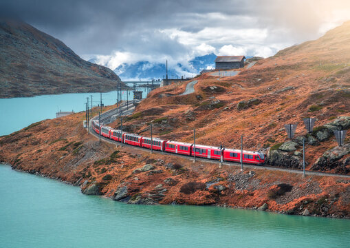 Aerial view of red train moving along turquoise lake in the Swiss Alps, rocky hills, dramatic cloudy sky in autumn alpine mountains. Top view of train in fall. Bernina Express, Switzerland. Railway