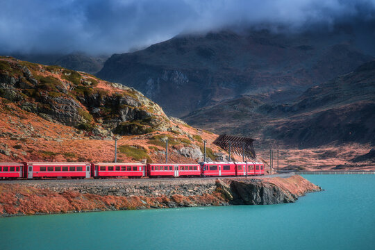 Aerial view of red train moving along the turquoise lake in the Swiss Alps, rocky hills, dramatic cloudy sky in alpine mountains in autumn. Top view of train in fall. Bernina Express, Switzerland