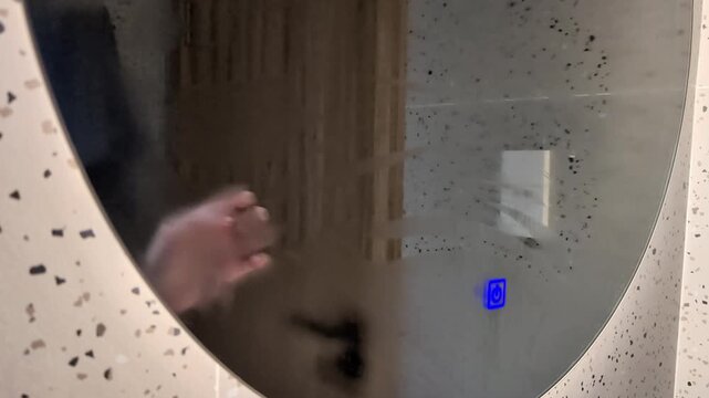 Close-up of a Caucasian female hand touching the sensor button of a round foggy bathroom mirror to turn on the built-in LED backlight. Then, the palm wipes away the condensation.