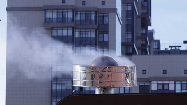 Shiny metallic vent cap atop building releases thick steam plume; urban backdrop with modern windows creates cool industrial mood