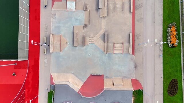 Straight-down drone view of an empty skatepark with concrete ramps, rails and stairs beside a bike lane, red paving and landscaped flower beds.