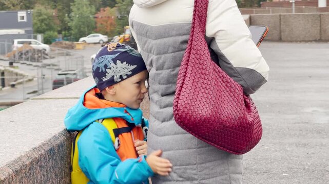 A curious boy leans affectionately against his mother on a granite embankment in Saint Petersburg. She holds a phone while they wait together. The sweet moment offers warmth and quiet comfort.