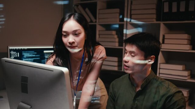 Two young Asian software developers brainstorming and working on a project, analyzing computer code on a desktop monitor in a modern office at night with lines of code projected on their faces