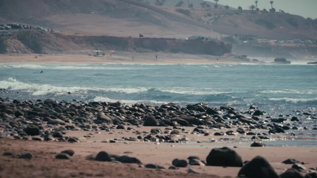 Waves roll toward the rocky shore of Banana Beach, Tamraght, Morocco, with the coastal hillside and parking area visible in the distance.