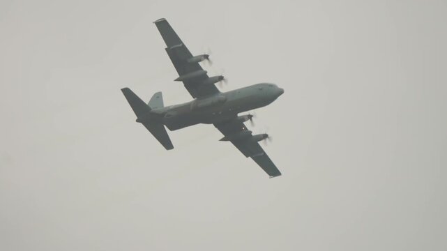 Low angle telephoto tracking shot of four engine military transport airplane flying across overcast sky past deciduous treetop during Falcon Leap airborne event near Oosterbeek.