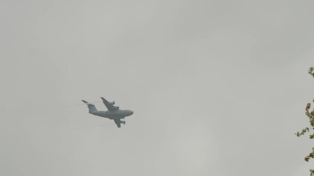 Telephoto view captures heavy turboprop transport plane disappearing behind leafy green and orange tree crown during Falcon Leap airborne exercise near Oosterbeek.