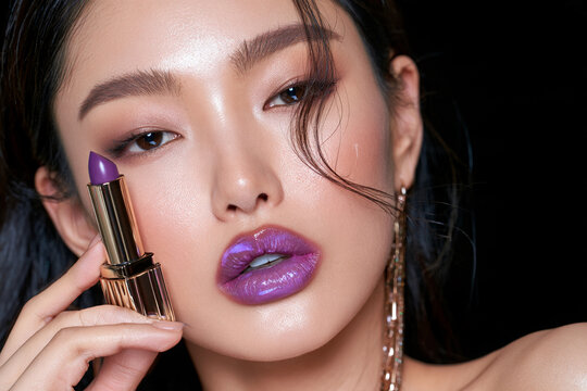 Asian woman poses with a tube of purple lipstick against black background. Cosmetics and make-up concept. Close-up fashion portrait for commercial poster