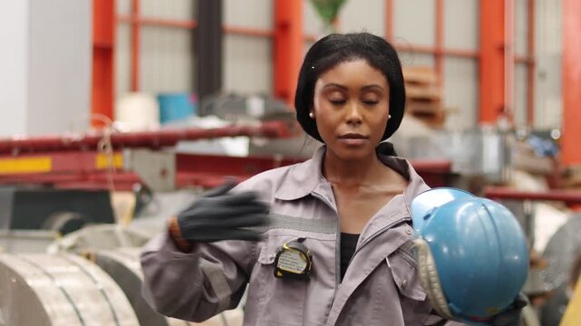African American worker with helmet in safety clothing wiping sweat off forehead while working in manufacturing factory, feel hot and tired. Exhausted female industrial engineer surrounded by machines