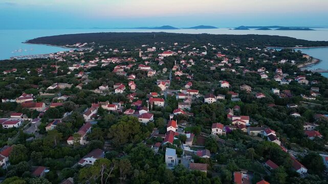 A breathtaking evening aerial drone panorama of old village on Silba Island, Croatia. The shot showcases the vibrant turquoise harbor, a docked ferry, and the charming coastal village stretching