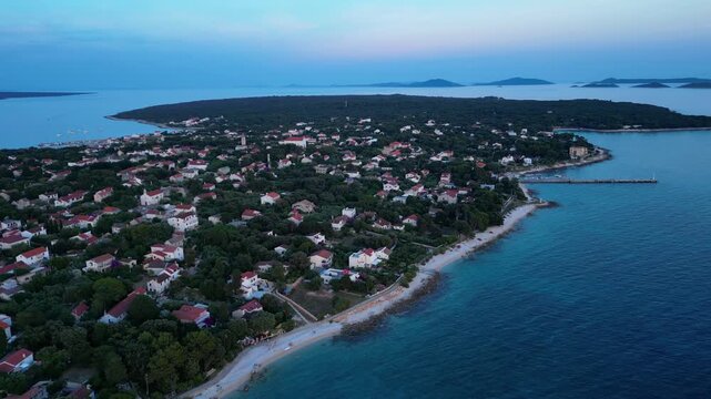 A breathtaking evening aerial drone panorama of old village on Silba Island, Croatia. The shot showcases the vibrant turquoise harbor, a docked ferry, and the charming coastal village stretching