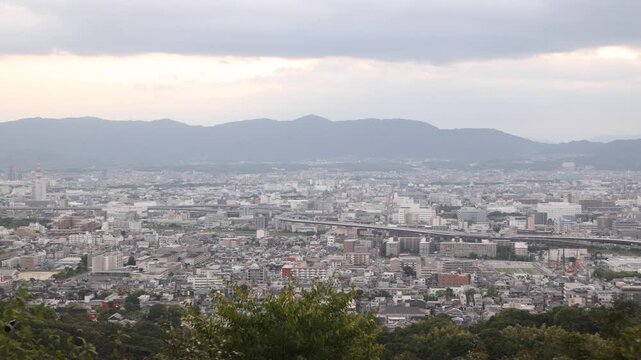panoramic view over Kyoto from the slopes of Mount Inari near Fushimi Inari Taisha, with the city extending toward distant hills beneath soft atmospheric light
