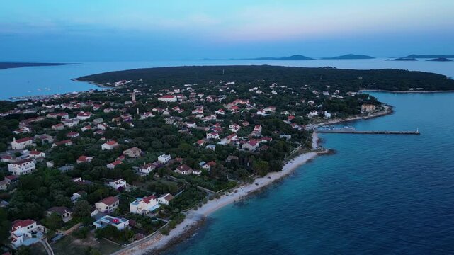 A breathtaking evening aerial drone panorama of old village on Silba Island, Croatia. The shot showcases the vibrant turquoise harbor, a docked ferry, and the charming coastal village stretching