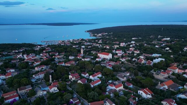 A breathtaking evening aerial drone panorama of old village on Silba Island, Croatia. The shot showcases the vibrant turquoise harbor, a docked ferry, and the charming coastal village stretching