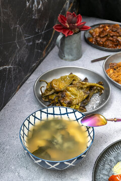 Miso-style broth with iridescent spoon beside grilled padron peppers, red flower accent, and shared dishes on marble counter.