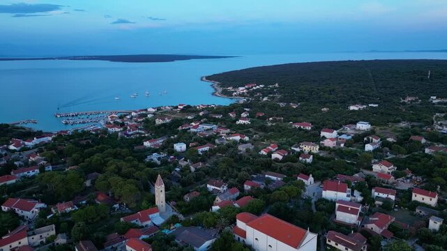 A breathtaking evening aerial drone panorama of old village on Silba Island, Croatia. The shot showcases the vibrant turquoise harbor, a docked ferry, and the charming coastal village stretching