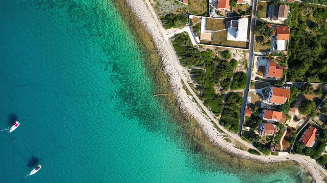 An aerial panoramic view of the tranquil turquoise bay on Silba Island, Croatia, featuring anchored sailboats along a lush, green Mediterranean coastline with charming red-roofed village houses under