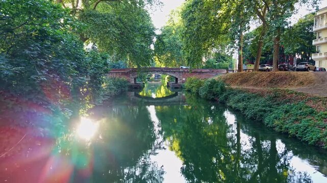 Low-altitude backward drone movement from a pedestrian brick bridge in Toulouse as cars pass along the shaded Garonne River green canal environment, Haute-Garonne, France