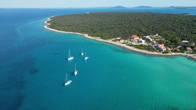 An aerial panoramic view of the tranquil turquoise bay on Silba Island, Croatia, featuring anchored sailboats along a lush, green Mediterranean coastline with charming red-roofed village houses under