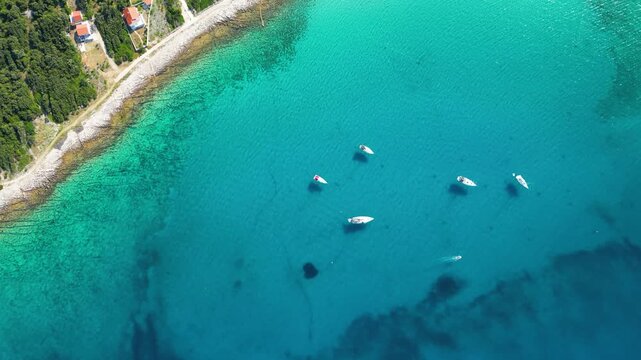 An aerial panoramic view of the tranquil turquoise bay on Silba Island, Croatia, featuring anchored sailboats along a lush, green Mediterranean coastline with charming red-roofed village houses under
