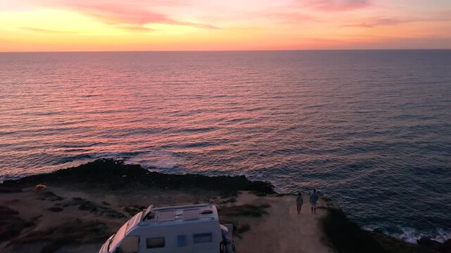  A couple watches the sunset in front of their camper van at La Frana Beach, Sardinia, Italy. Waves lap against the rocky shore as visitors enjoy the tranquility at sunset